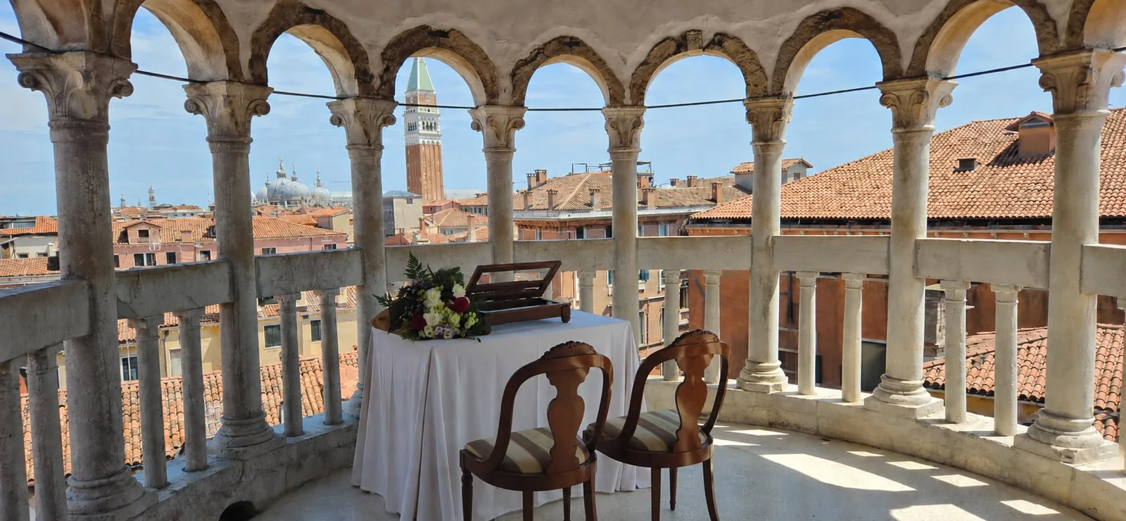 A ceremony table dressed in white with a bouquet and two wooden chairs on a curved open terrace, overlooking the rooftops of Venice
