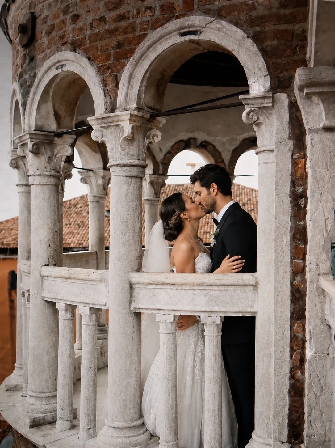 A bride and groom kissing on a historic Venetian loggia, framed by stone columns and arches, with terracotta rooftops behind them