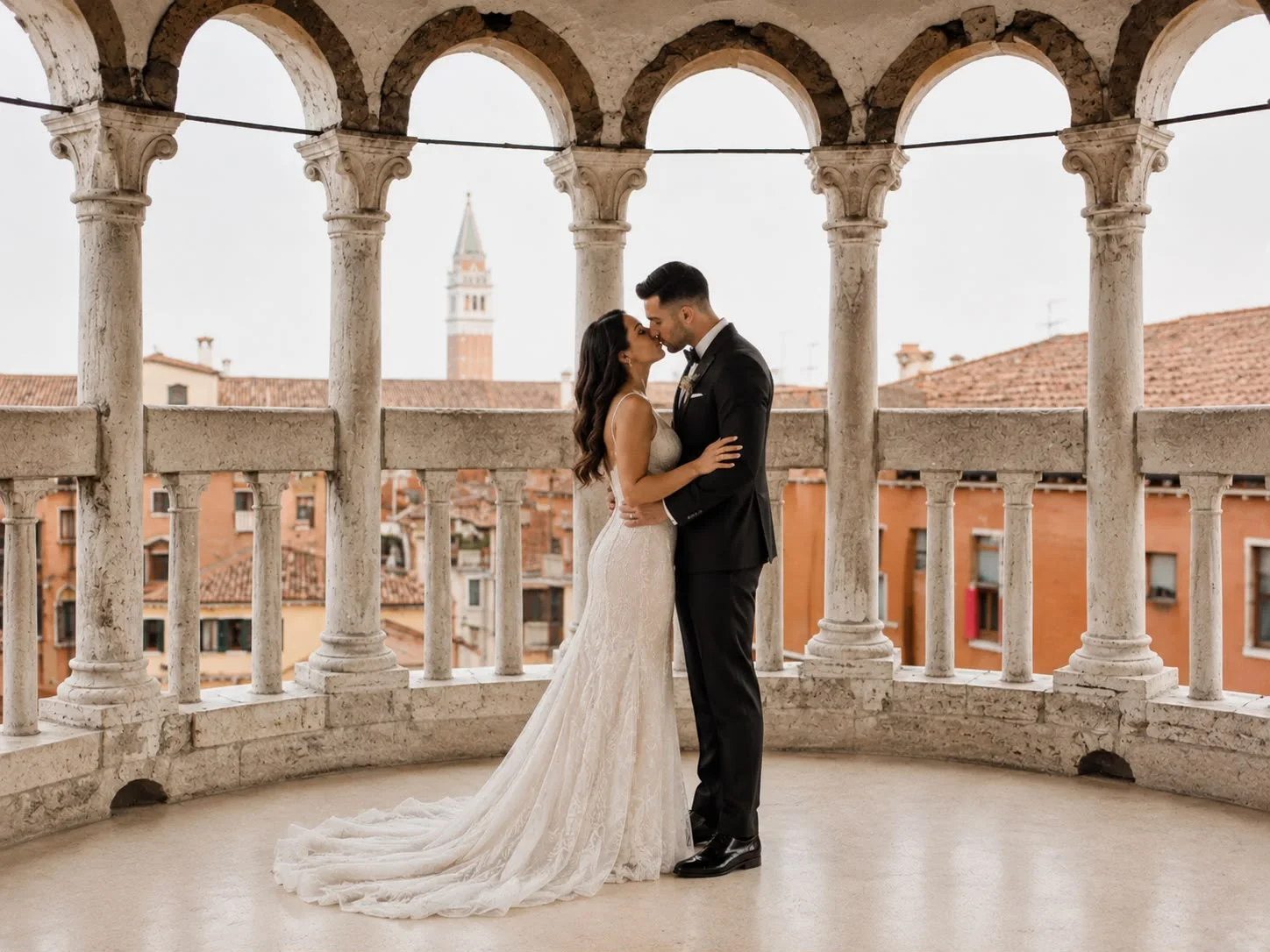 A couple kissing on a historic Venetian loggia of stone columns and arches, with tiled rooftops and a bell tower behind them