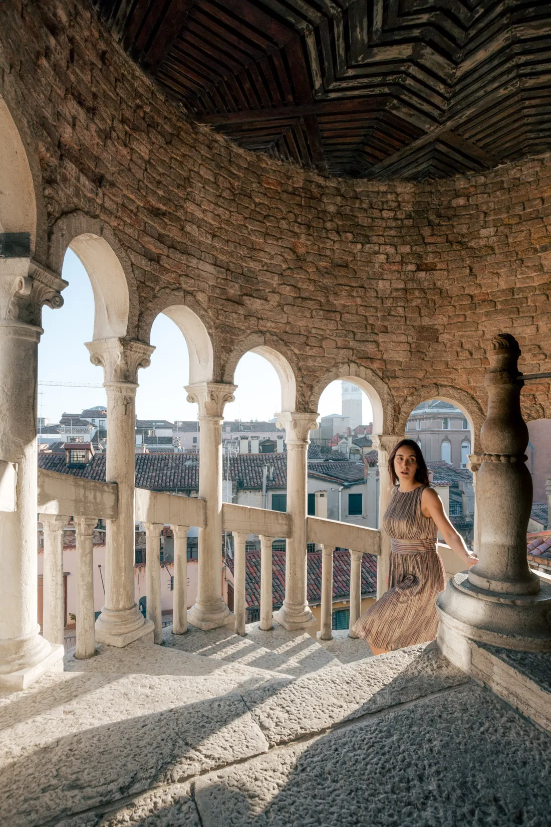 A woman in a light evening dress in the loggia of stone arches, looking out over Venice