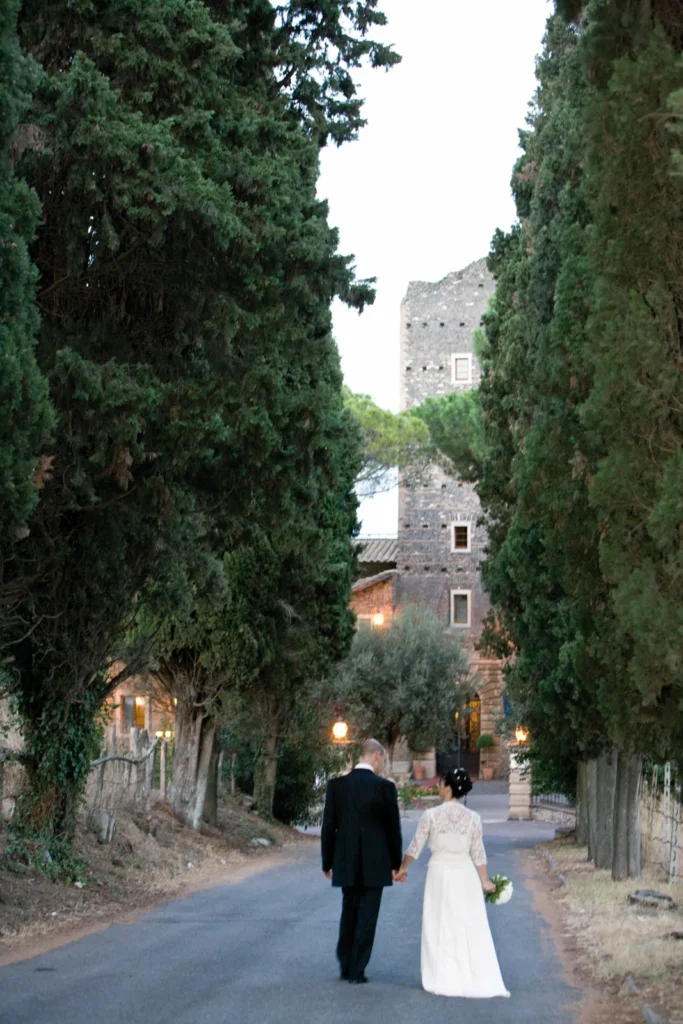 A bride and groom walking hand in hand along a cypress-lined avenue towards the castle tower