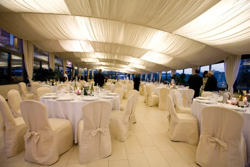 A bright glass-walled pavilion set for a wedding dinner, with draped ceiling and round tables