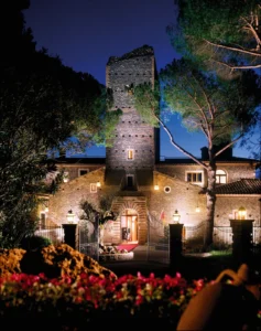 A medieval castle tower lit up at night above a garden of red flowers, just outside Rome