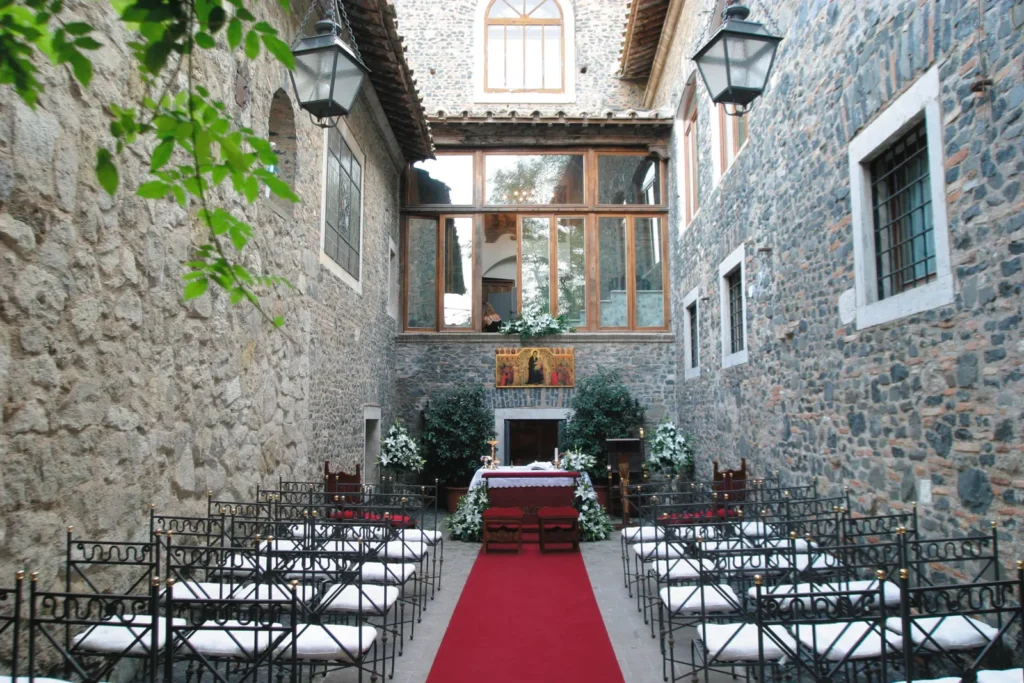 A stone cloister set for a wedding ceremony, with a red carpet, white chairs and flowers
