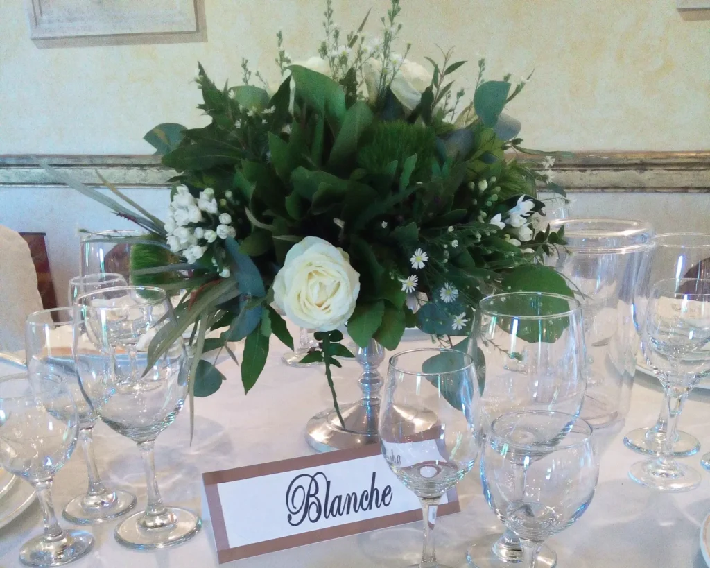 A wedding table with a centerpiece of white roses and greenery, crystal glasses and a name card