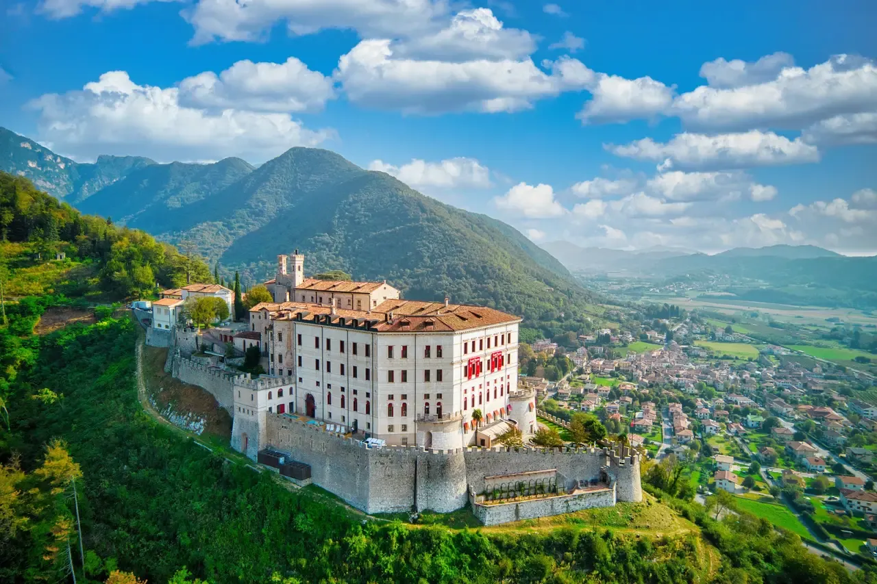 Aerial view of a large Italian castle on a green hilltop, with the valley and mountains of the Veneto around it
