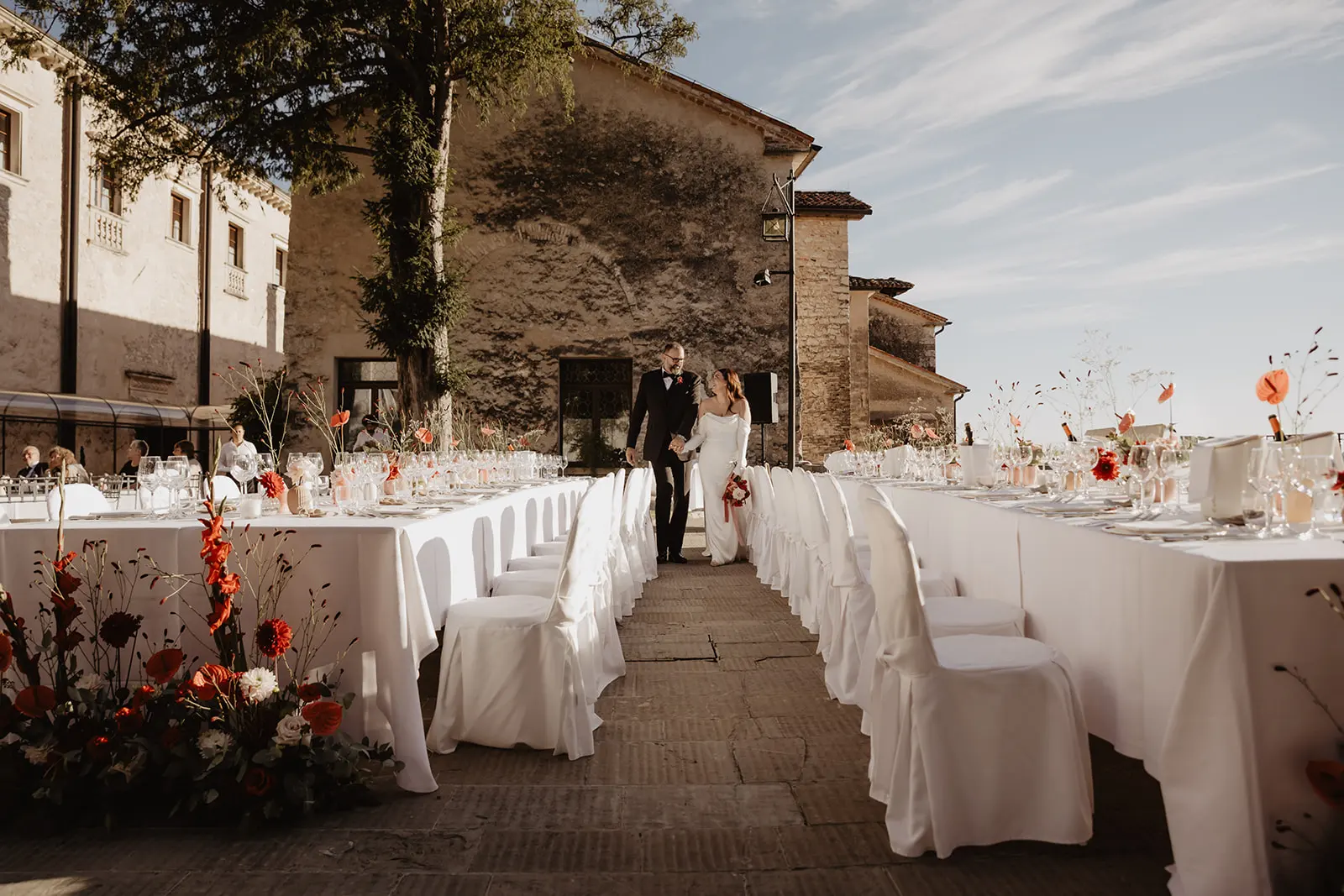 Long wedding tables dressed in white on a castle terrace, with flowers and a couple walking between them