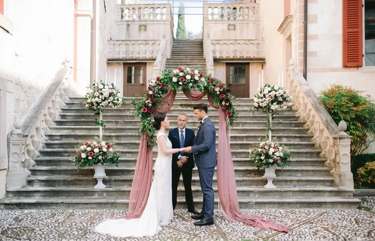 A couple during a symbolic wedding ceremony under a floral arch at the foot of a stone staircase