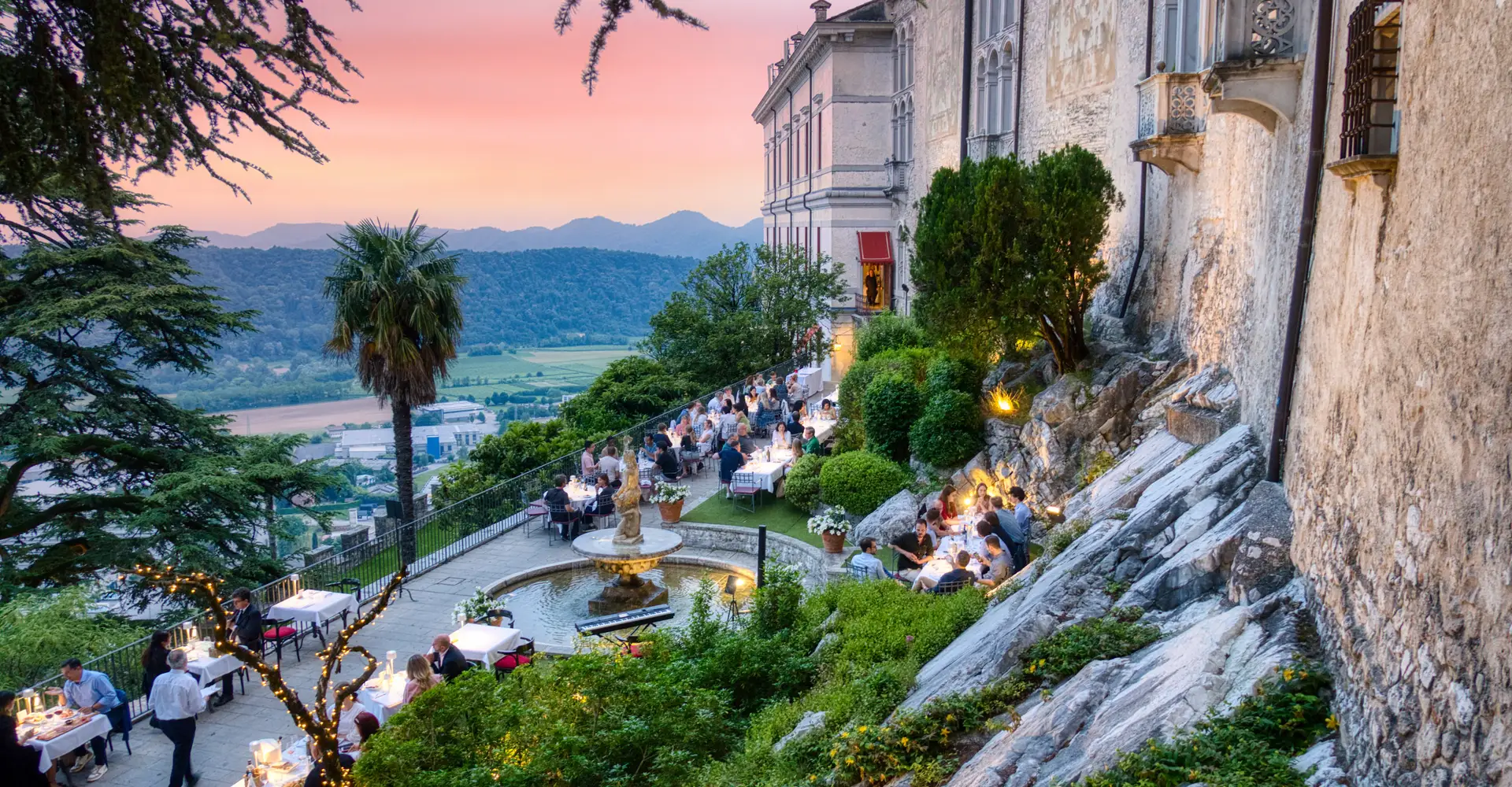 Guests dining on the terraces of a hilltop castle at sunset, with a palm tree, gardens and the mountains of the Veneto behind