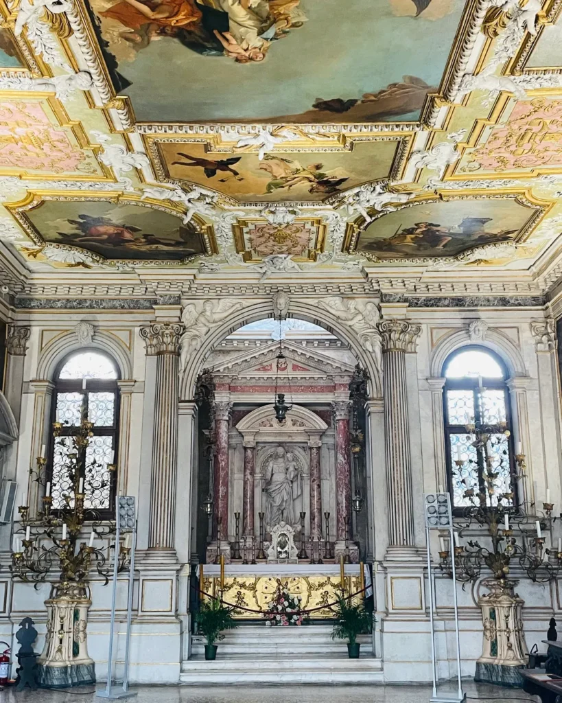 El altar de mármol de la Sala Capitular de un palacio barroco veneciano, entre dos ventanales, bajo un techo pintado y dorado