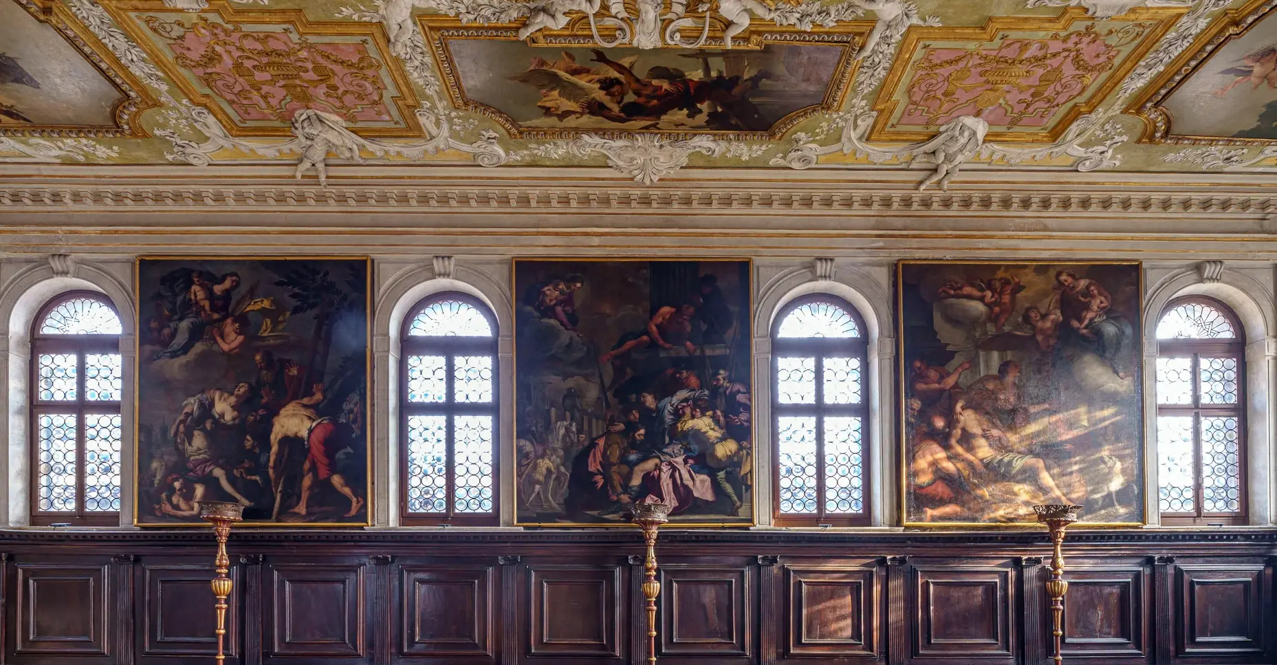 The Chapter Hall of a Baroque Venetian palace, with a painted ceiling, large canvases and carved wooden benches along the walls