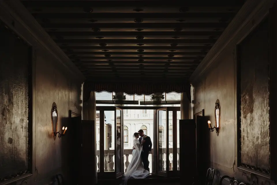 A bride and groom embracing at the open balcony doors at the end of a long palace hall