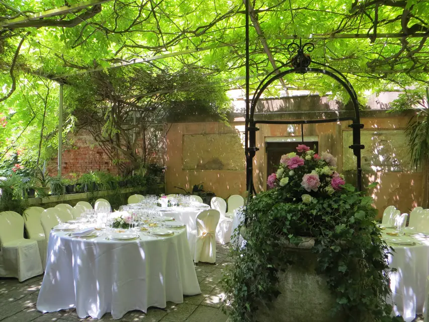 Round tables dressed in white for a dinner under a green pergola, with a pink flower arrangement