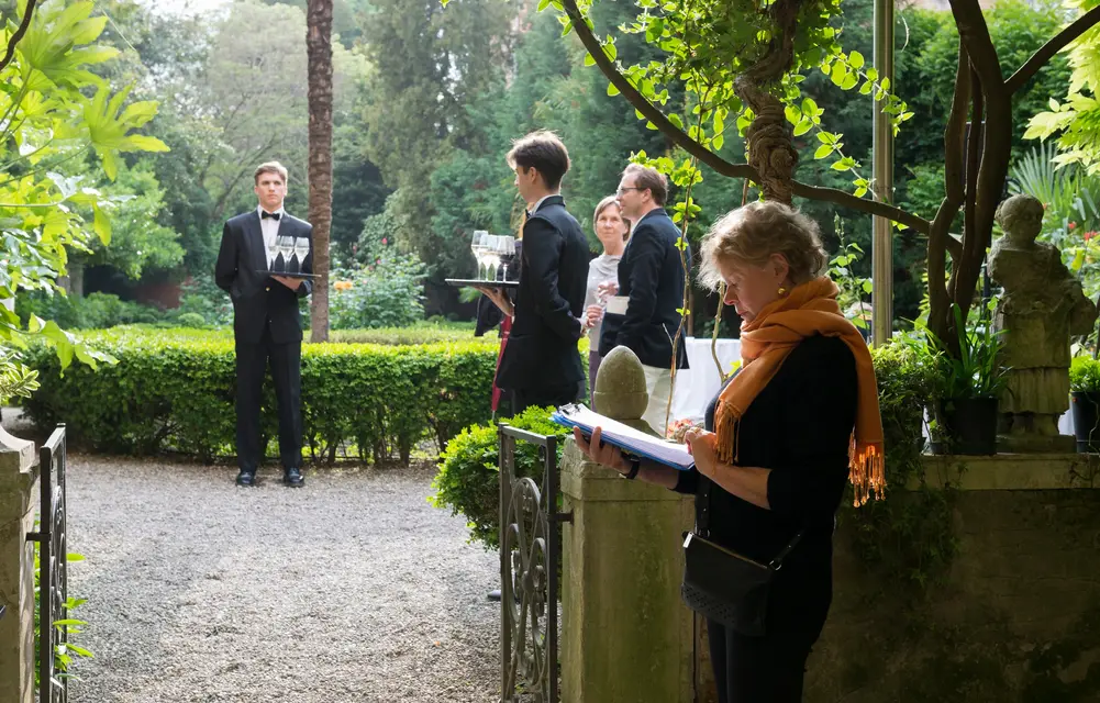 Waiters with trays of prosecco glasses in the garden, beside a woman reading notes