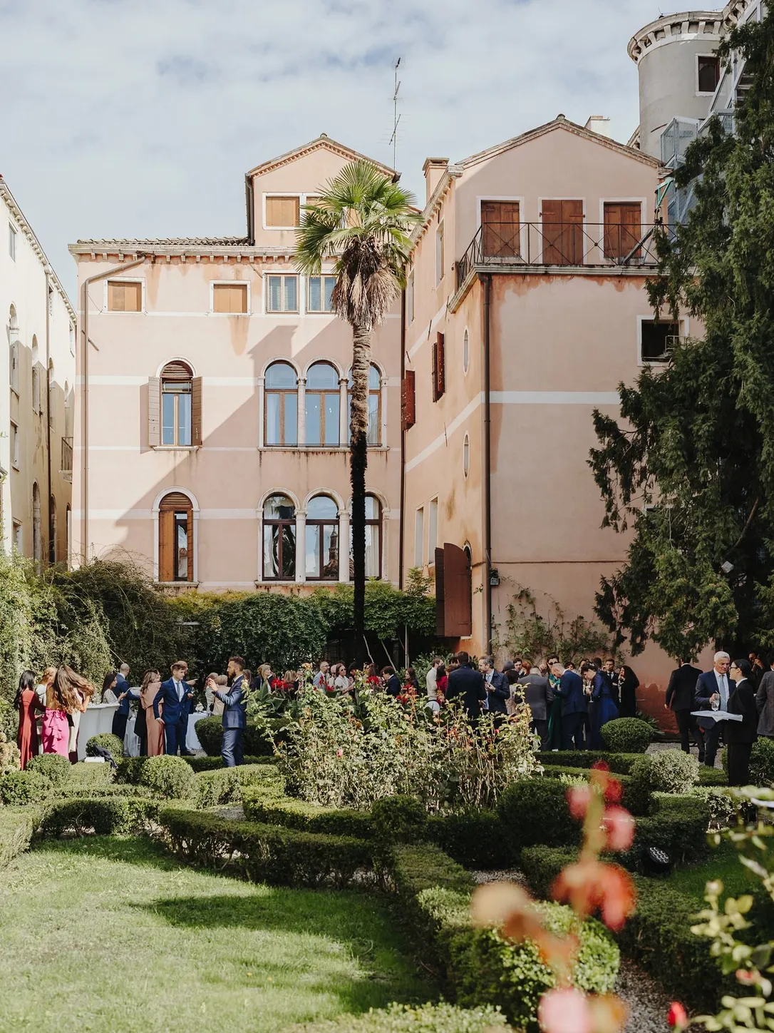 Guests gathered for a ceremony in the palace garden, in front of a pink Venetian facade with arched windows and a palm tree