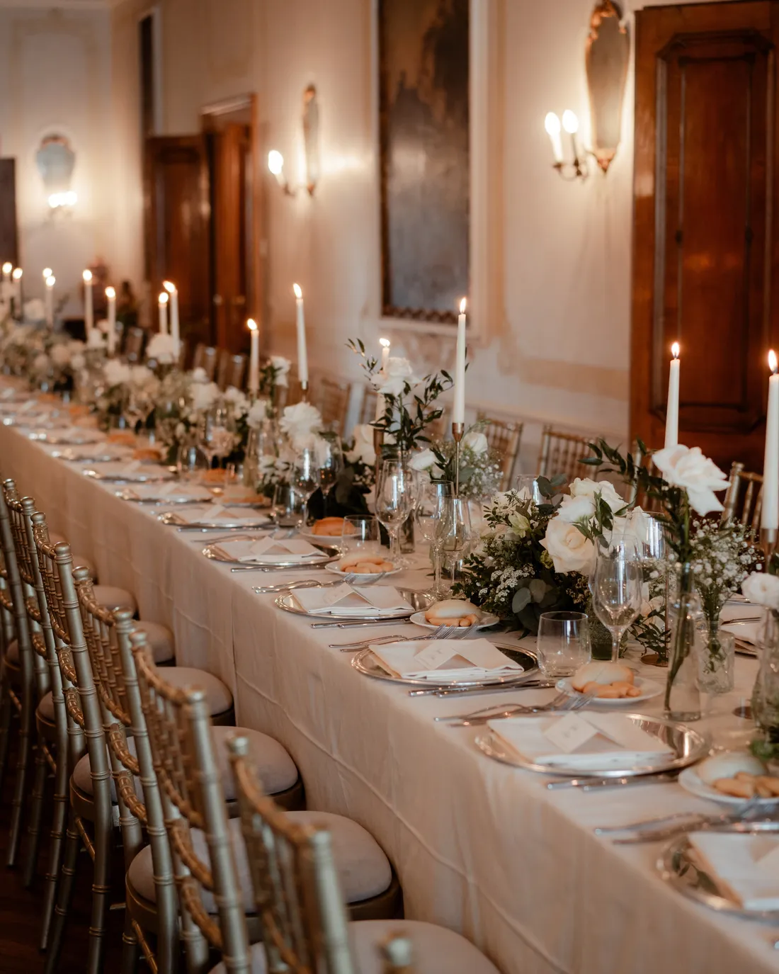 A long candlelit wedding dinner table with white flowers along a frescoed palace hall