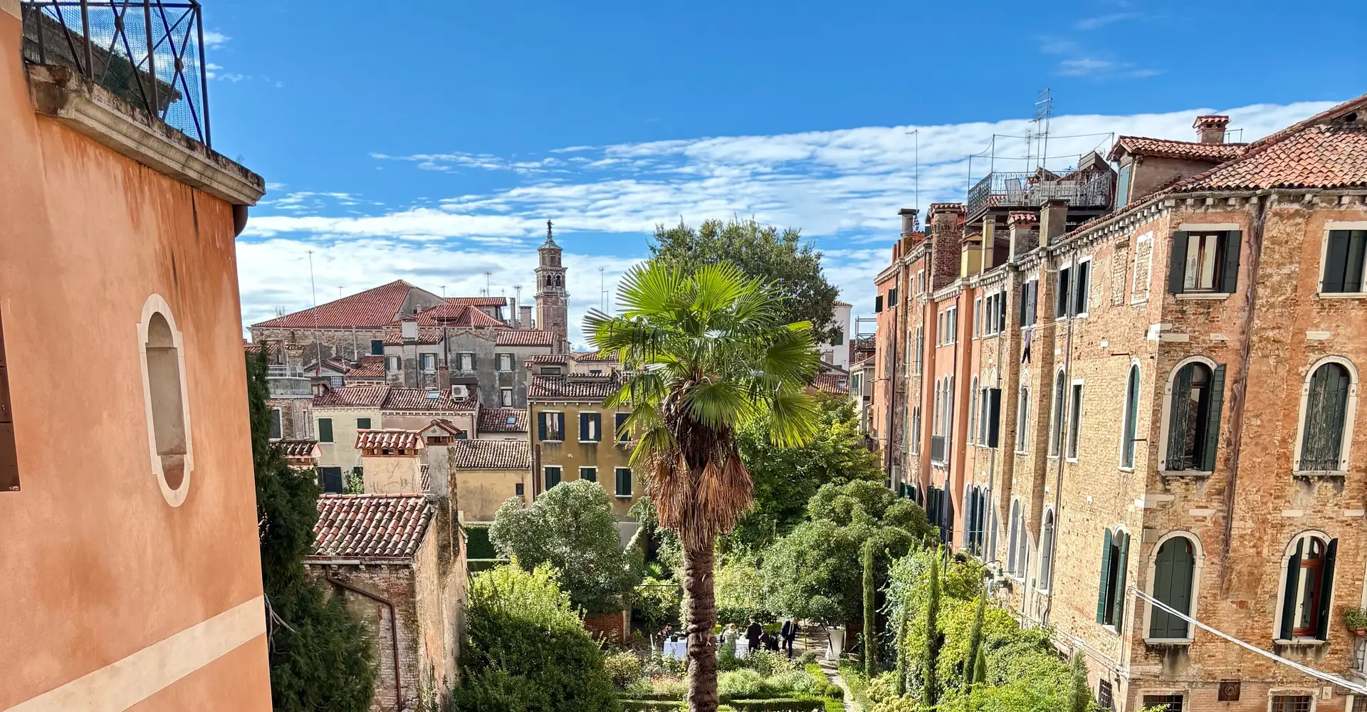 A tall palm tree in an Italian-style garden hidden between Venetian palaces, with a bell tower and blue sky behind